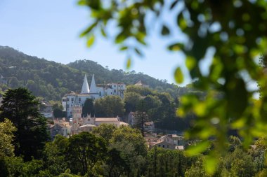 Sintra Ulusal Sarayı manzarası ön planda ağaç yaprakları ile çerçevelenmiş yemyeşil tepelerle çevrili. İkiz bacaları olan tarihi beyaz saray Portekiz 'in kültürel simgesidir..