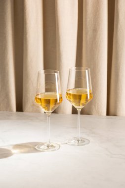 Arka planda bej perdeleri olan mermer bir masada iki bardak beyaz şarap, ılık ışıkta yumuşak gölgeler bırakır..