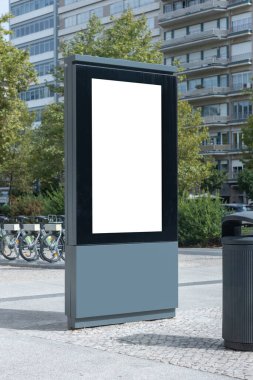 Urban advertising panel with a blank space for announcements, located in a square in Lisbon, Portugal. In the background, there is a modern residential building, green trees, and public shared bicycles, creating a contemporary city atmosphere.