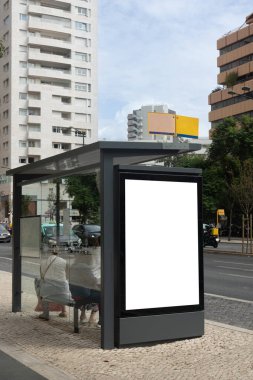 Bus stop with a large blank advertising billboard on a busy avenue, surrounded by tall modern buildings and people waiting inside the shelter. Lisbon Portugal