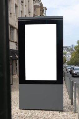 Lisbon, Portugal - Vertical blank billboard with white mockup space on a sidewalk, surrounded by city buildings and parked cars on a cobblestone street.