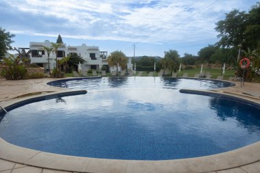 Luxury resort pool in Algarve, Portugal, surrounded by tropical gardens and white Mediterranean-style villas. The calm blue water reflects the partly cloudy sky, creating a relaxing holiday atmosphere.