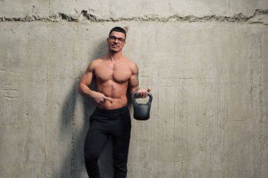 Gözlüklü genç ve güçlü bir adam beton bir duvarın arkasında elinde bir kettlebell tutuyor.
