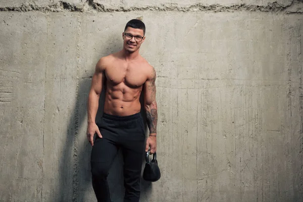 Gözlüklü genç ve güçlü bir adam beton bir duvarın arkasında elinde bir kettlebell tutuyor.
