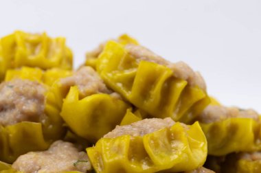 Shumai olarak bilinen buharda pişirilmiş Çin hamur köftelerinin yakın plan çekimi, canlı sarı bir ambalaj kağıdı ve temiz beyaz bir arkaplan üzerine kurulu tuzlu bir et dolgusu içeriyor..