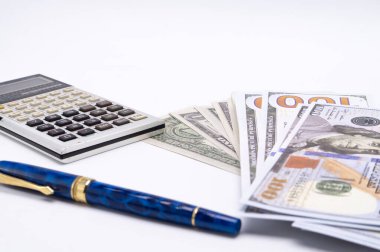 Bir hesap makinesi, mavi bir kalem ve beyaz bir yüzeye yığılmış yüz dolarlık banknotlar, finans ve parayı temsil ediyor..