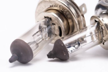 İki halojen araba farı ampulünün detaylı çekimi. Temiz cam, gümüş metal taban ve beyaz zemin üzerine kurulmuş koyu gri bir uç..