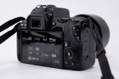 Siyah bir DSLR kameranın beyaz bir yüzeyde duran mercek ve kayışla yakın çekimi. Kamera görüntüsü görünür, çeşitli ayarları gösterir.