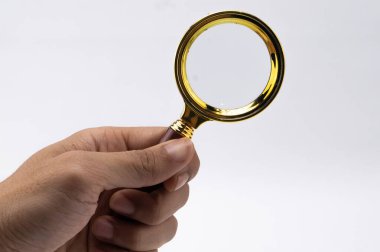 Altın çerçeveli bir büyüteçle temiz beyaz bir arkaplan üzerinde duran bir elin yakın çekimi, ayrıntıları vurgulamak ve incelemek.