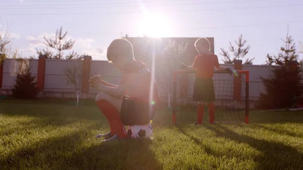 Niño pequeño está sentado en la pelota y otro en el gol de fútbol en el césped en el patio trasero — Foto de Stock