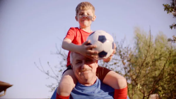 小さな男の子は彼の祖父の首の上に座って、彼の手の中でサッカーボールを保持している — ストック写真