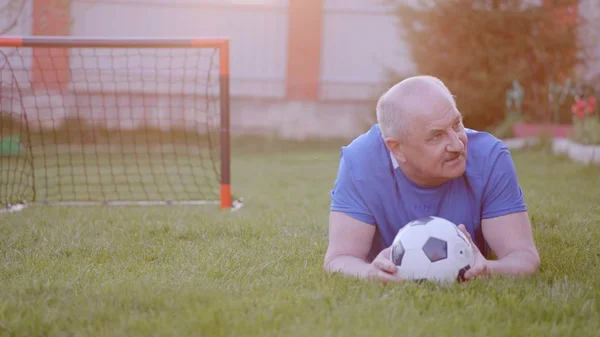 Starý muž na fotbalovém uniformě leží na trávníku s fotbalovým míčkem — Stock fotografie