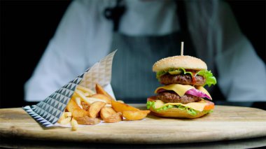 Şef lezzetli hamburger ve patates kızartması siyah arka plan üzerine ahşap tepsi üzerinde hizmet veren
