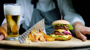 Şef lezzetli hamburger, kızarmış patates ve bira siyah arka plan üzerine ahşap tepsi üzerinde hizmet veren
