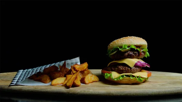 Close up of appetizing burger with double cheese and golden french fries on the wooden tray. Black background — Stock Photo, Image