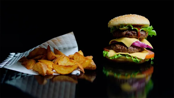 Kentang goreng dan burger dengan keju ganda dan sayuran segar di latar belakang hitam dan refleksi pada permukaan kaca — Stok Foto