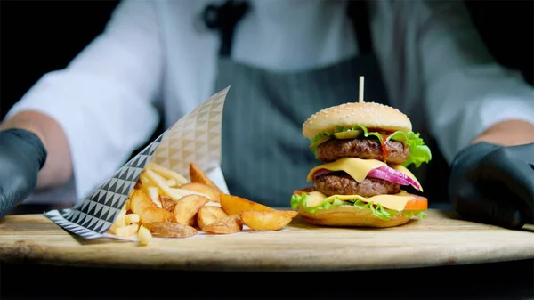 Şef lezzetli hamburger ve patates kızartması siyah arka plan üzerine ahşap tepsi üzerinde hizmet veren