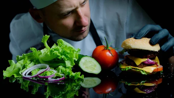 Chef terminando la sabrosa hamburguesa con carne de res, queso y verduras frescas. Fondo negro para comercial —  Fotos de Stock