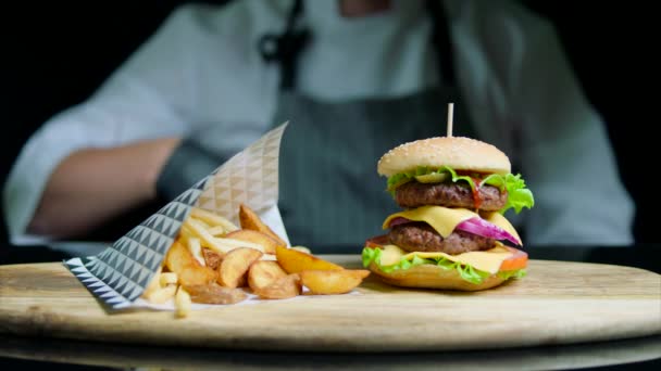 Chef está mostrando el pulgar fuera de foco y conjunto de sabrosa hamburguesa, papas fritas, cerveza sobre fondo negro — Vídeos de Stock