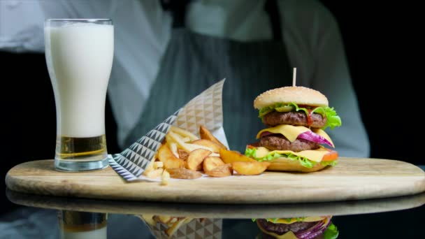 Chef che serve un gustoso hamburger, patatine fritte, versando birra nel bicchiere su un vassoio di legno — Video Stock