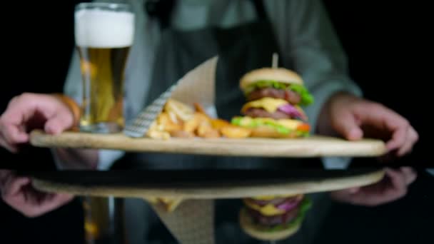 Koch serviert leckere Burger, Pommes, Bier auf dem hölzernen Tablett auf schwarzem Hintergrund — Stockvideo