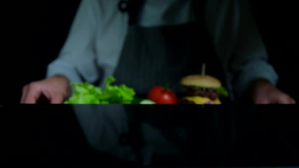 Färska grönsaker och välsmakande hamburgare med ost och nötkött kotletter. Kock på bakgrunden hamnar ur fokus — Stockvideo