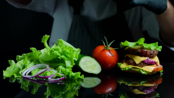 Chef finissant le burger salé avec du bœuf, du fromage et des légumes. Fond noir pour commercial — Video