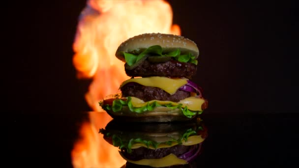 Вкусный горячий бургер на поверхности черного зеркала в огне. Черный фон для рекламы — стоковое видео