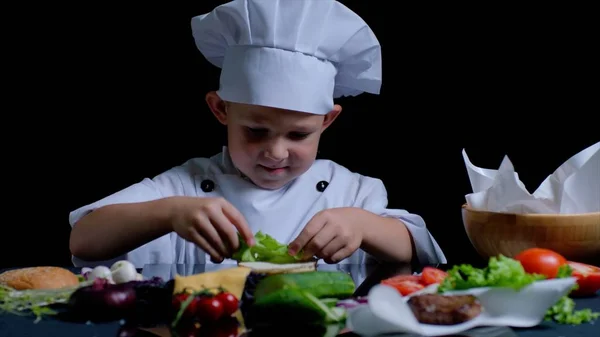 Маленький мальчик готовит бургер на кухне, на нем костюм шеф-повара. — стоковое фото