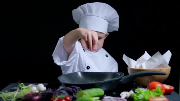 Маленький мальчик добавляет соль в блюдо в костюме шеф-повара и кепке. Черный фон для рекламы — стоковое видео