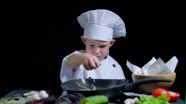 Menino está adicionando um sal ao prato, vestindo terno de chefs e boné. Fundo preto para comercial — Vídeo de Stock