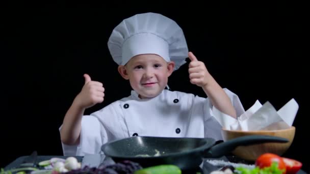 Menino alegre está dançando e mostrando os polegares enquanto cozinha. Ele usa terno e boné de chefs — Vídeo de Stock