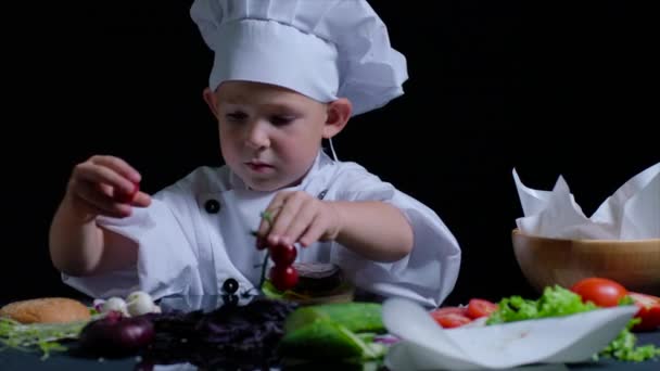 Приятный мальчик готовит бургер в костюме шеф-повара и кепке. Черный фон для рекламы — стоковое видео