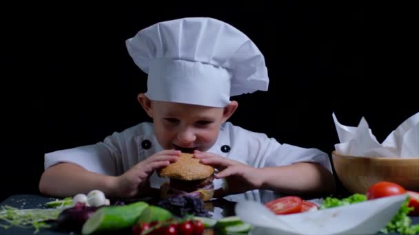 Aranyos fiú főzés közben eszik egy hamburgert. Visel, szakácsok suit és a sapka. — Stock videók