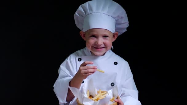 Chico sonriente con traje de chef come papas fritas en el fondo negro — Vídeo de stock