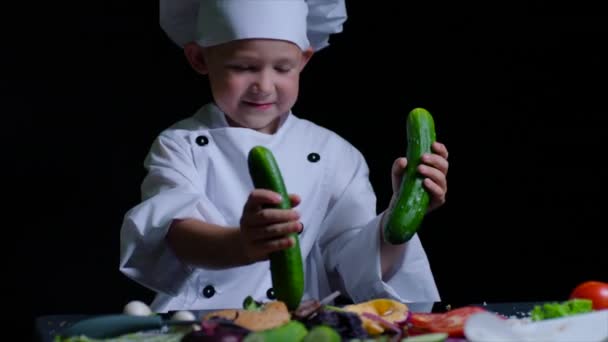 Joli garçon portant des chefs costume joue avec les concombres à la table de cuisson. Fond noir — Video