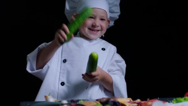 Merry boy con traje de chef juega con pepinos en la mesa de cocina. Fondo negro — Vídeo de stock