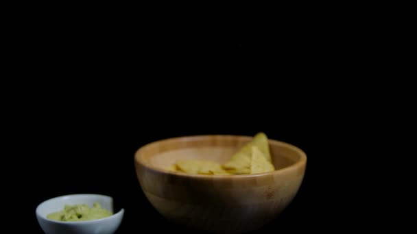 Crispy nachos is falling into the wooden bawl. Spicy guacomole sause at the foreground — Stock Video