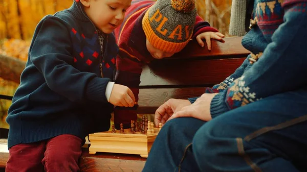 Los hermanos pequeños están jugando ajedrez con papá en el banco en el parque de otoño — Foto de Stock