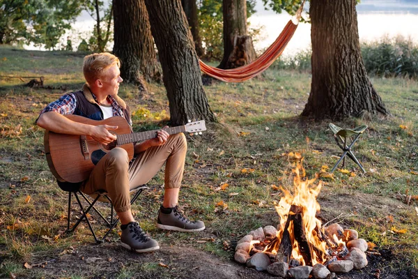 Genç adam zammı üzerinde kamp şenlik ateşi yakınındaki gitar çalmak