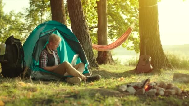 Jeune homme travaillant à l'ordinateur portable pendant le camping avec des tentes au coucher du soleil — Video