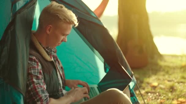 Hombre joven está trabajando en el portátil mientras está sentado en la tienda de campaña en el camping durante el senderismo — Vídeo de stock