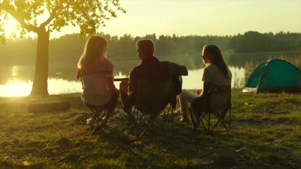 Backshot de amigos disfrutando de la puesta de sol en el lago en el camping con tiendas de campaña — Vídeos de Stock
