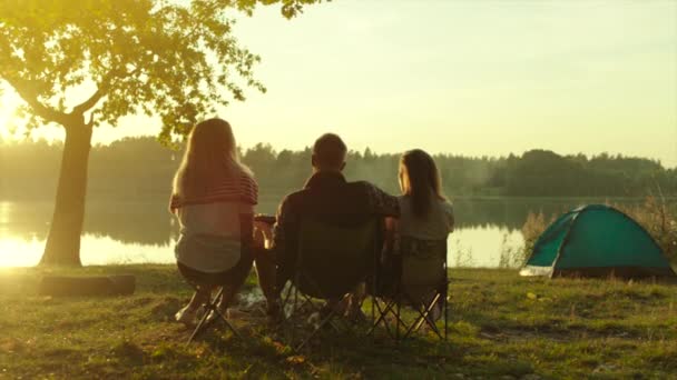 Backshot av vänner njuter av solnedgången vid sjön på camping med tält — Stockvideo