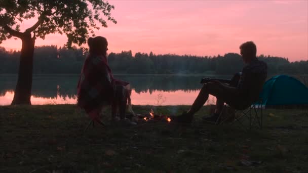 Pareja joven está disfrutando de la puesta de sol en el senderismo, tocando la guitarra cerca de la hoguera en el camping — Vídeos de Stock