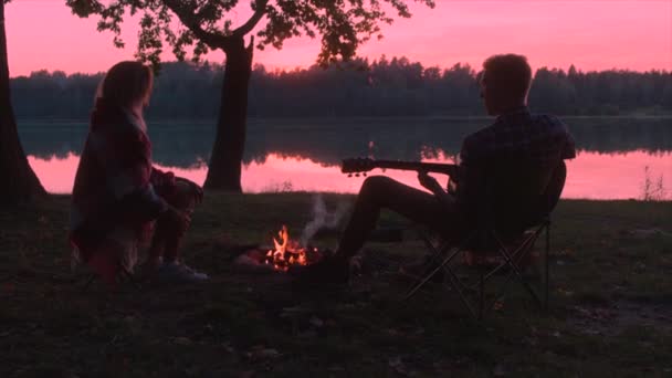 Młody człowiek jest gra na gitarze do jego dziewczyna siedząc przy ognisku o zachodzie słońca — Wideo stockowe