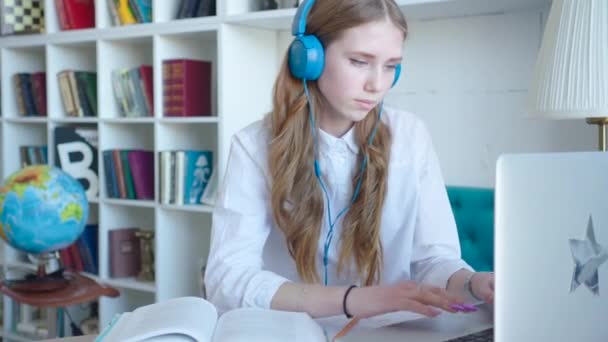 Hermosa chica adolescente que se prepara para el examen utilizando el ordenador portátil en la biblioteca, usando auriculares — Vídeo de stock