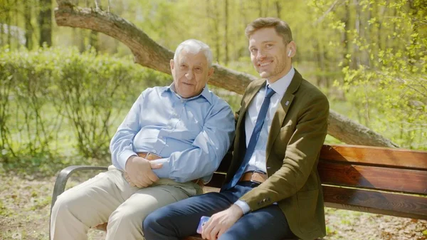 El viejo y su hijo sonríen, sentados en el banco del parque el día de verano. Imagen De Stock