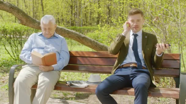 Старий читає книгу в парку, а бізнесмен перериває його гучною розмовою — стокове відео