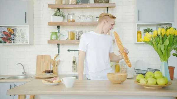 Young man is goofing around and singing with bread instead of microphone at the kitchen